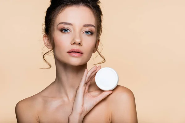 Pretty model with naked shoulders holding container with cosmetic cream isolated on beige — Stock Photo