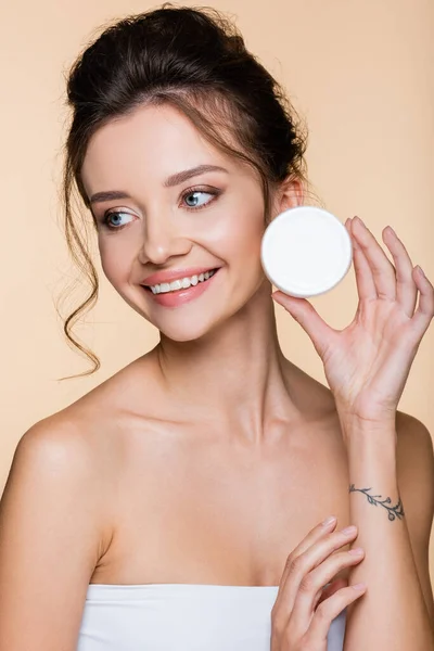 Modelo alegre segurando recipiente com creme cosmético isolado no bege — Fotografia de Stock