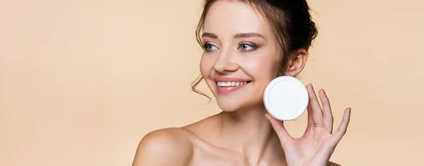 Pretty model with naked shoulders holding cosmetic cream isolated on beige, banner — Stock Photo