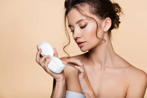 Mujer morena sosteniendo contenedor con crema cosmética aislada en beige — Stock Photo