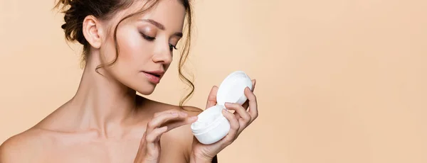 Young model with naked shoulders holding cosmetic cream and cap isolated on beige, banner — Stock Photo
