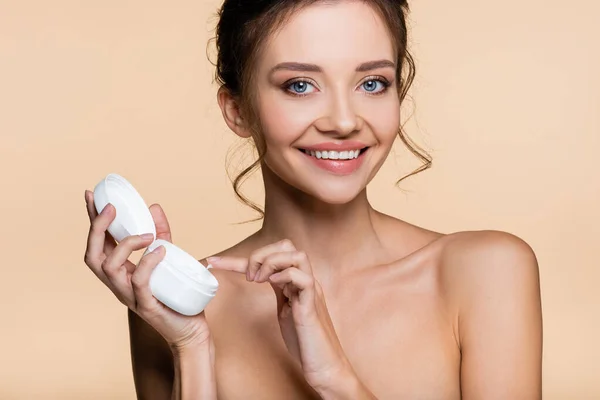 Mujer sonriente con hombros desnudos sosteniendo crema cosmética y mirando a la cámara aislada en beige — Stock Photo