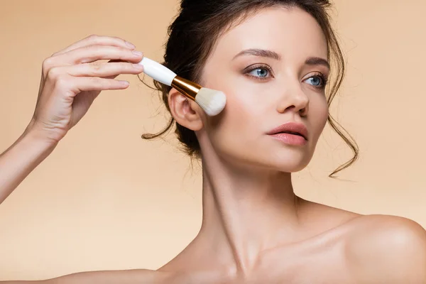 Young model with naked shoulders holding cosmetic brush near cheek isolated on beige — Stock Photo