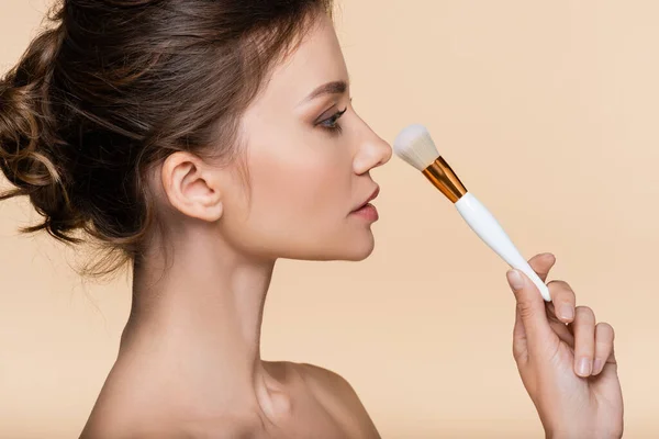 Vista laterale della donna bruna che tiene la spazzola cosmetica vicino al naso isolato sul beige — Foto stock