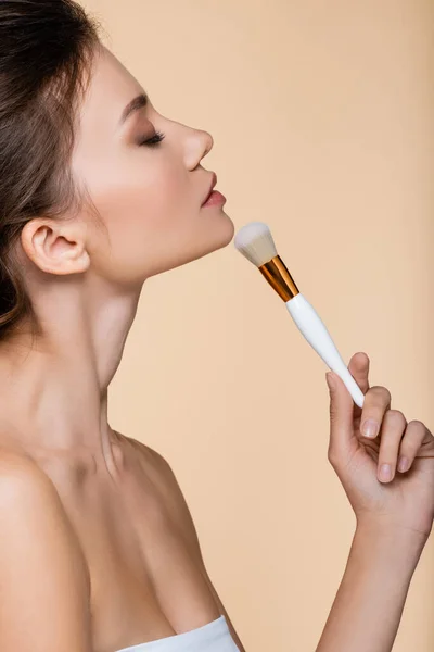 Side view of pretty model with closed eye holding cosmetic brush near chin isolated on beige — Stock Photo