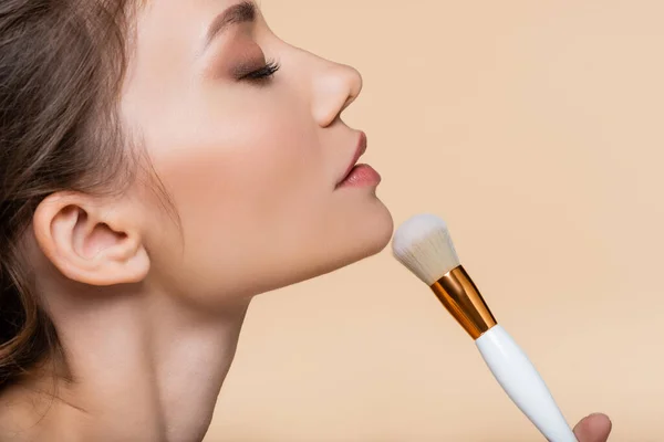 Side view of young woman holding cosmetic brush near chin isolated on beige — Stock Photo