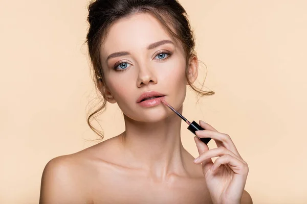 Young woman with naked shoulders applying lip gloss isolated on beige — Stock Photo