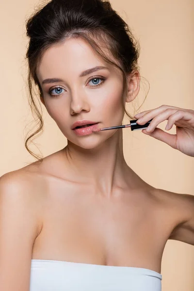 Mujer joven en top blanco aplicando brillo de labios y mirando a la cámara aislada en beige - foto de stock