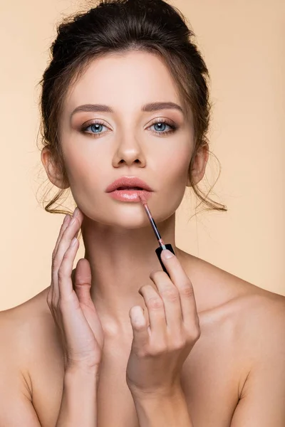 Young model with naked shoulders applying lip gloss and touching face isolated on beige — Stock Photo