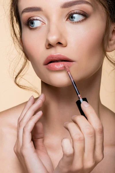 Portrait of pretty woman applying lip gloss and looking away isolated on beige — Stock Photo