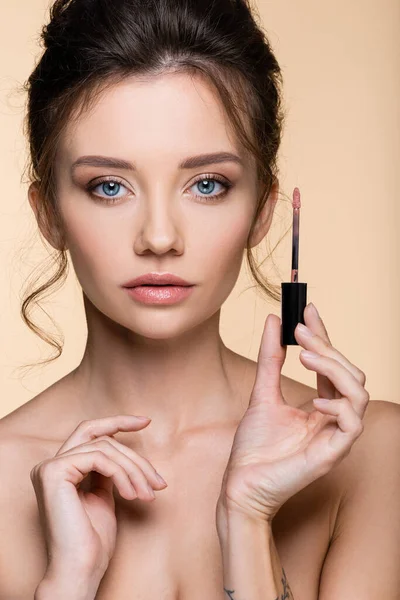 Retrato de modelo bonito sosteniendo cepillo de brillo de labios y mirando a la cámara aislada en beige - foto de stock