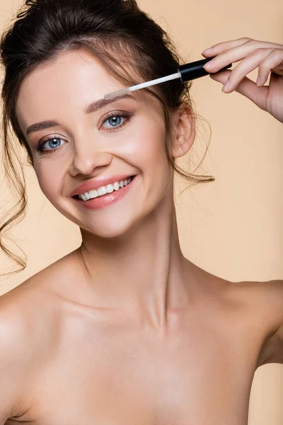 Smiling model with naked shoulders holding brush of eyebrow gel isolated on beige — Stock Photo