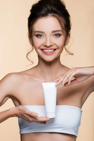 Smiling brunette woman in white top holding cosmetic lotion isolated on beige — Stock Photo