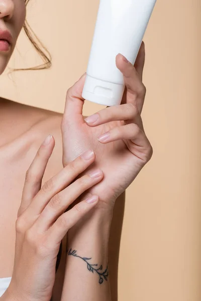 Cropped view of tattooed woman holding tube with cosmetic lotion isolated on beige — Stock Photo