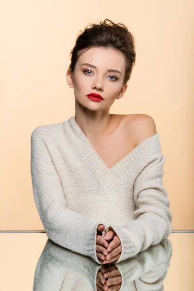 Young model in warm sweater looking at camera near mirror isolated on beige — Stock Photo