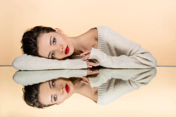 Modelo elegante em suéter deitado no espelho isolado no bege — Fotografia de Stock
