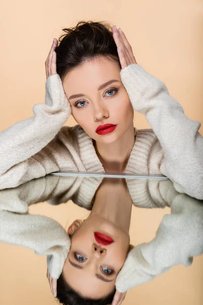 Elegante modelo morena tocando el pelo cerca del espejo aislado en beige - foto de stock