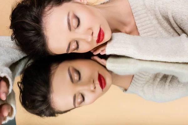 Modelo elegante em suéter fechando os olhos enquanto deitado no espelho isolado no bege — Fotografia de Stock