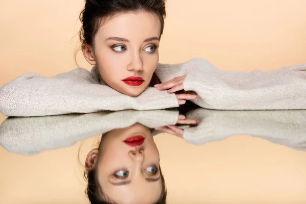 Modelo bonito com lábios vermelhos vestindo suéter e olhando para longe perto do espelho isolado no bege — Fotografia de Stock