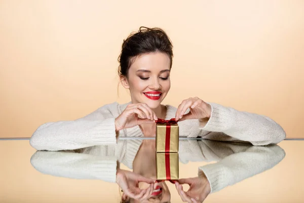 Donna sorridente in maglione caldo con fiocco di regalo su specchio isolato su beige — Foto stock