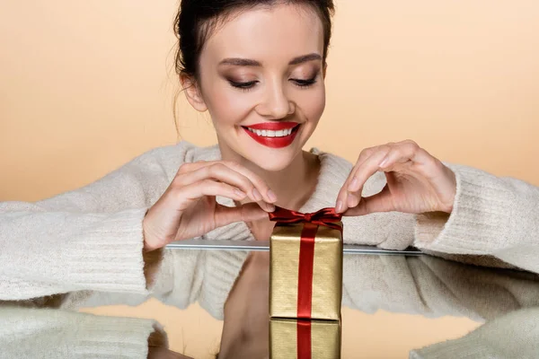 Femme positive avec lèvres rouges tenant arc de boîte cadeau d'or près du miroir isolé sur beige — Photo de stock