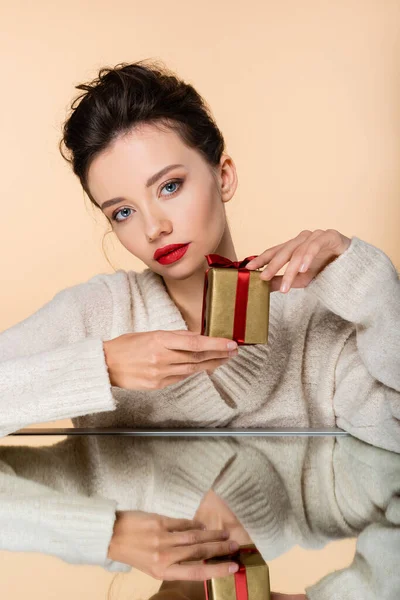 Stilvolle Frau im Pullover hält Geschenk in der Nähe Spiegel isoliert auf beige — Stockfoto