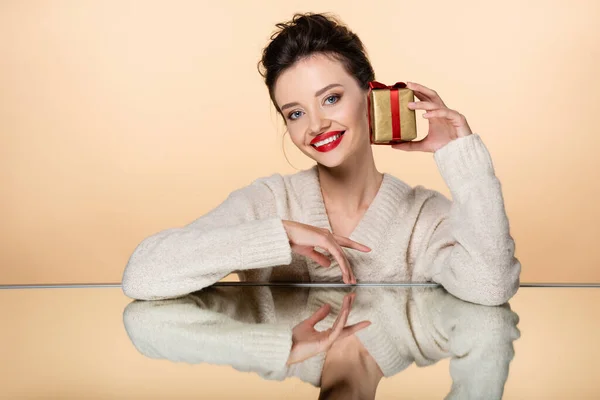 Jovem modelo feliz em suéter segurando caixa de presente dourado perto do espelho isolado no bege — Fotografia de Stock