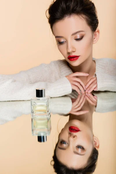 Elegante giovane donna guardando bottiglia di profumo su specchio isolato su beige — Foto stock