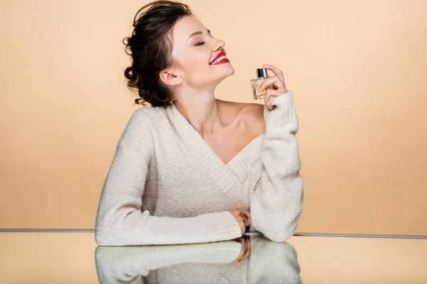 Mujer con estilo feliz en suéter con perfume cerca del espejo aislado en beige - foto de stock