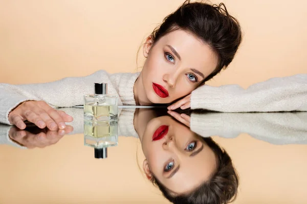 Modelo morena con labios rojos mirando a la cámara cerca de perfume en espejo aislado en beige - foto de stock