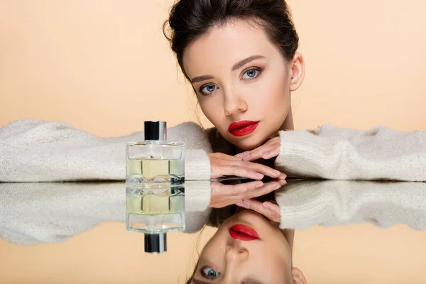 Stilvolle junge Frau in warmem Pullover posiert in der Nähe von Parfümflasche auf Spiegel isoliert auf beige — Stockfoto