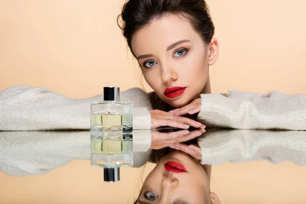 Mujer joven con estilo en suéter cálido posando cerca de la botella de perfume en el espejo aislado en beige - foto de stock