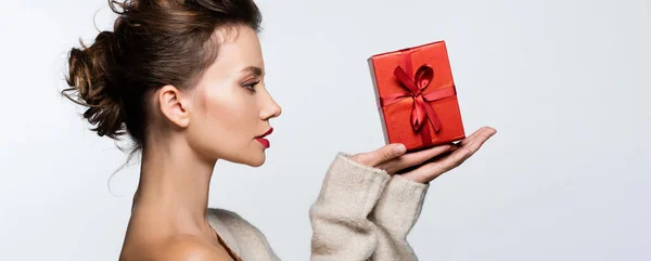 Vista lateral de modelo morena en suéter mirando caja de regalo con cinta aislada en blanco, banner - foto de stock