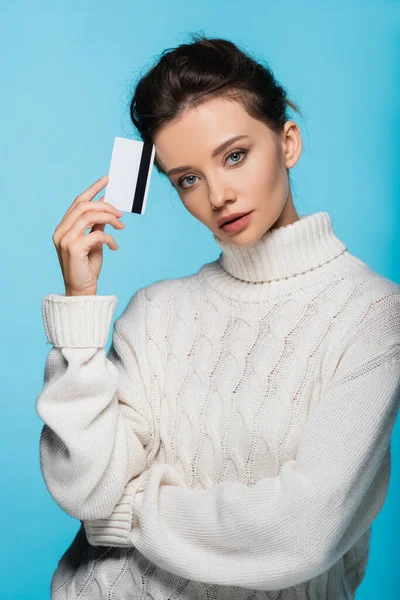 Brünette Frau im gemütlichen Pullover mit Kreditkarte und Blick in die Kamera isoliert auf blau — Stockfoto