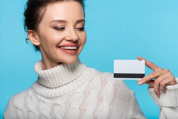 Smiling woman in knitted sweater looking at credit card isolated on blue — Stock Photo