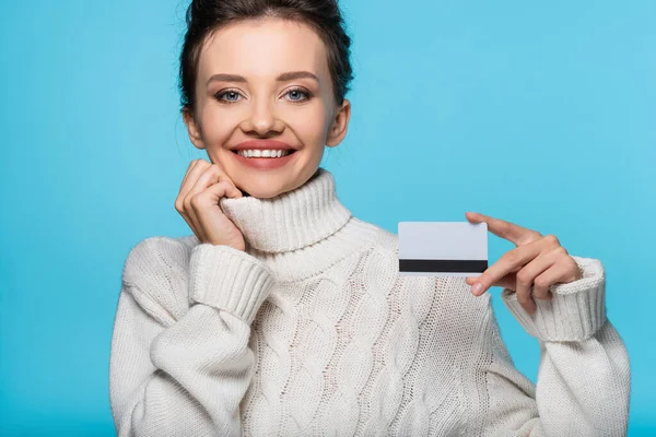 Mujer positiva en suéter caliente con tarjeta de crédito y mirando a la cámara aislada en azul - foto de stock