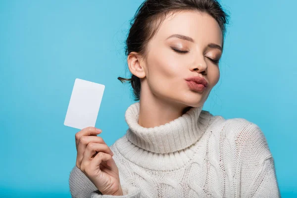 Mujer morena en suéter acogedor soplado beso de aire y la celebración de la tarjeta en blanco aislado en azul - foto de stock