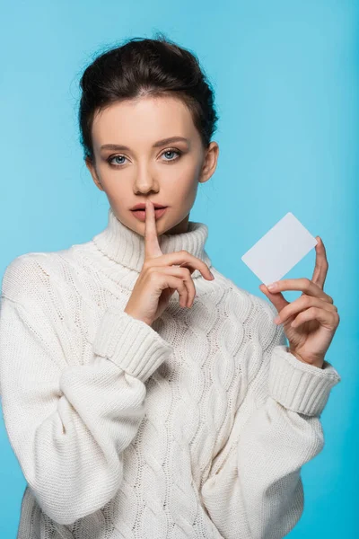 Modelo jovem em camisola de malha segurando cartão em branco e mostrando gesto secreto isolado em azul — Fotografia de Stock