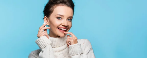 Smiling woman in knitted sweater holding wireless earphone isolated on blue, banner — Stock Photo