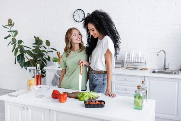 Happy woman looking at curly african american girlfriend holding knife in kitchen — Stock Photo