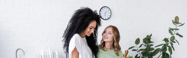 Happy woman looking at curly african american girlfriend in kitchen, banner — Stock Photo