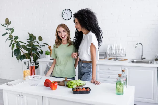 Mulher americana africana feliz olhando para namorada loira na cozinha — Fotografia de Stock