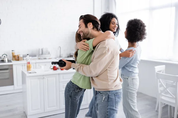 Glücklicher Mann mit Flasche Wein umarmt Frau neben glücklichen afrikanisch-amerikanischen Freunden zu Hause — Stockfoto