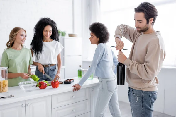 Hombre apertura botella de vino cerca interracial las mujeres en casa - foto de stock