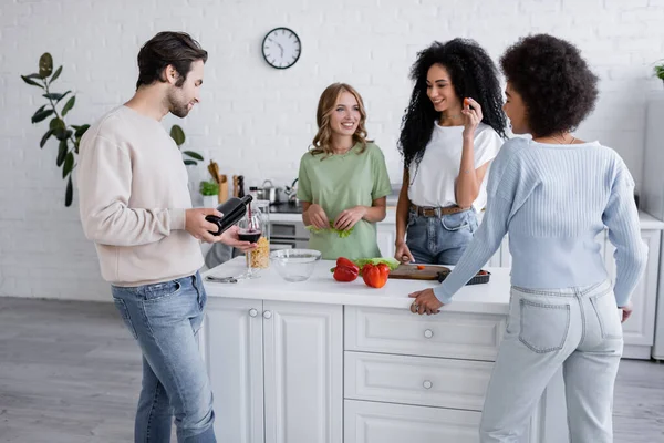 Homem derramando vinho tinto em vidro perto de mulheres inter-raciais na cozinha — Fotografia de Stock