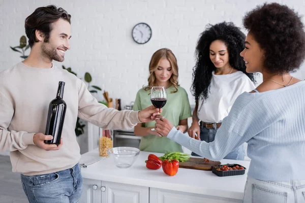 Uomo allegro che dà un bicchiere di vino alla donna afro-americana vicino agli amici in cucina — Foto stock