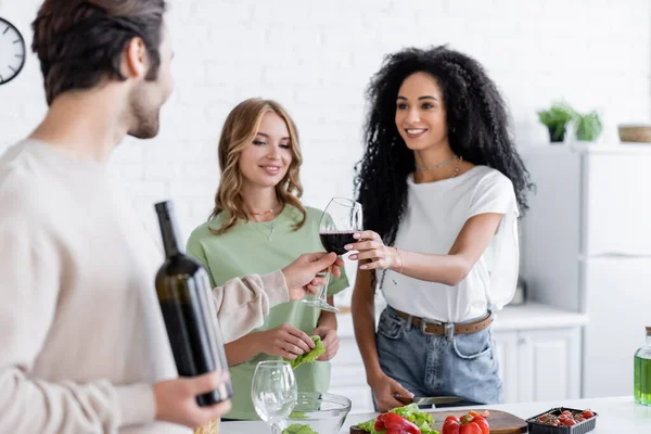 Homem desfocado dando copo de vinho para mulher americana africana alegre perto de amigo loiro na cozinha — Fotografia de Stock