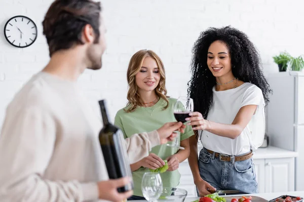 Hombre borroso dando copa de vino a la mujer afroamericana complacida cerca de amigo rubio en la cocina - foto de stock