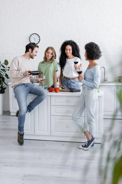 Volle Länge des Mannes gießt Rotwein in Glas neben glücklichen interrassischen Frauen in der Küche — Stockfoto