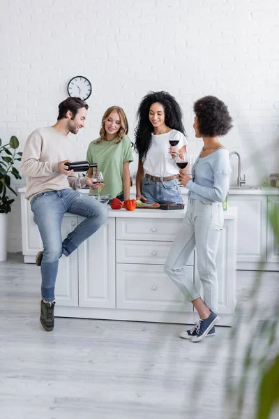 Longitud completa del hombre verter vino tinto en vidrio cerca de las mujeres interracial feliz en la cocina - foto de stock